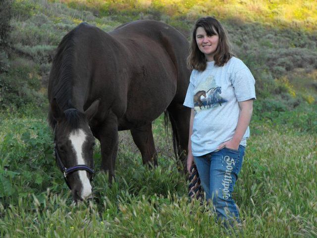 Woman smiling next to brown horse in grassy field with hillside background